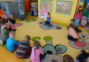 Dziewczynka opowiada dinozaurach leżących przed nią na dywanie, reszta dzieci siedzi w półkolu i zaciekawieniem słucha koleżanki.